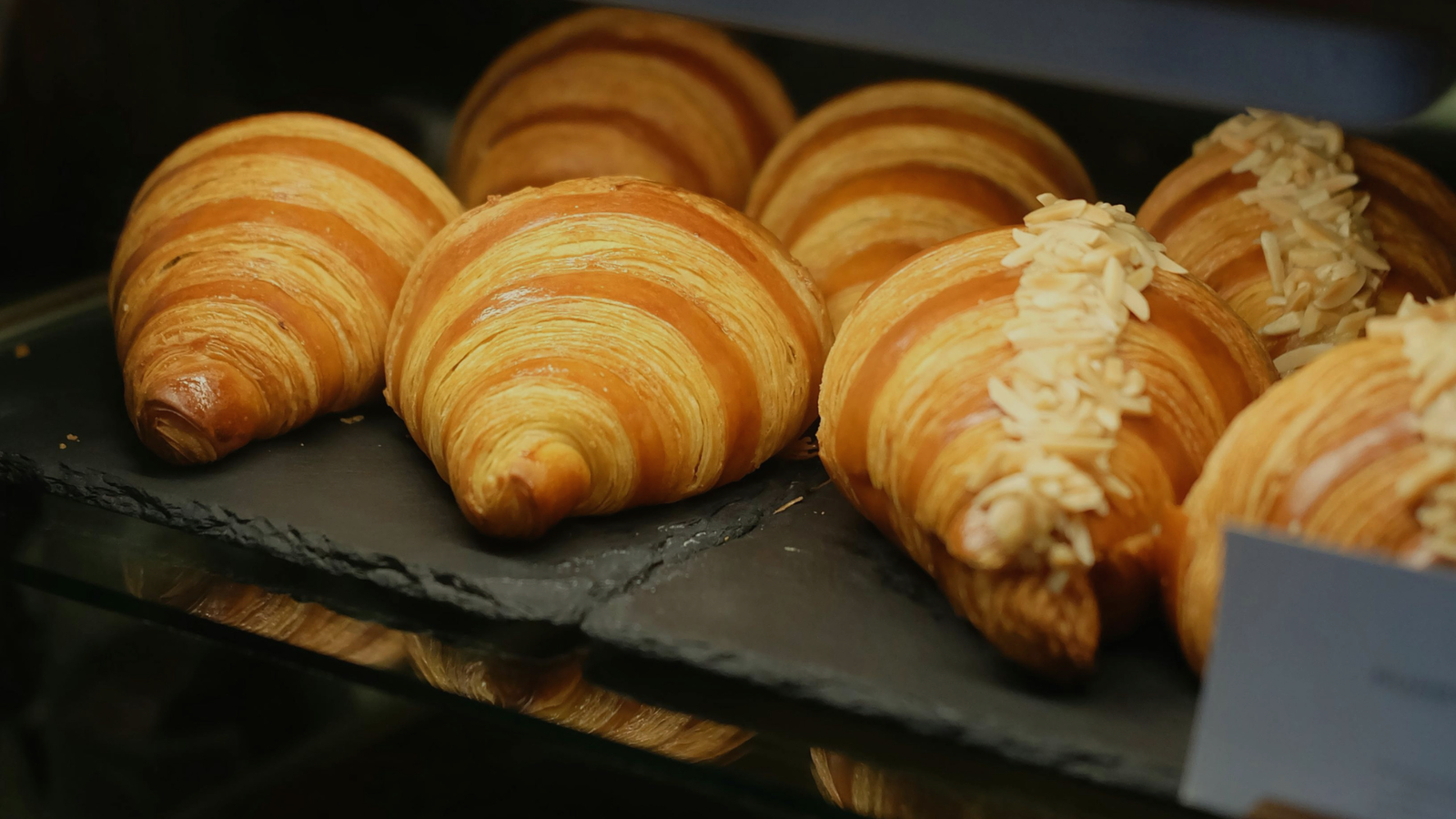 A close-up shot of several golden-brown, flaky croissants displayed on a black slate tray in a bakery case.